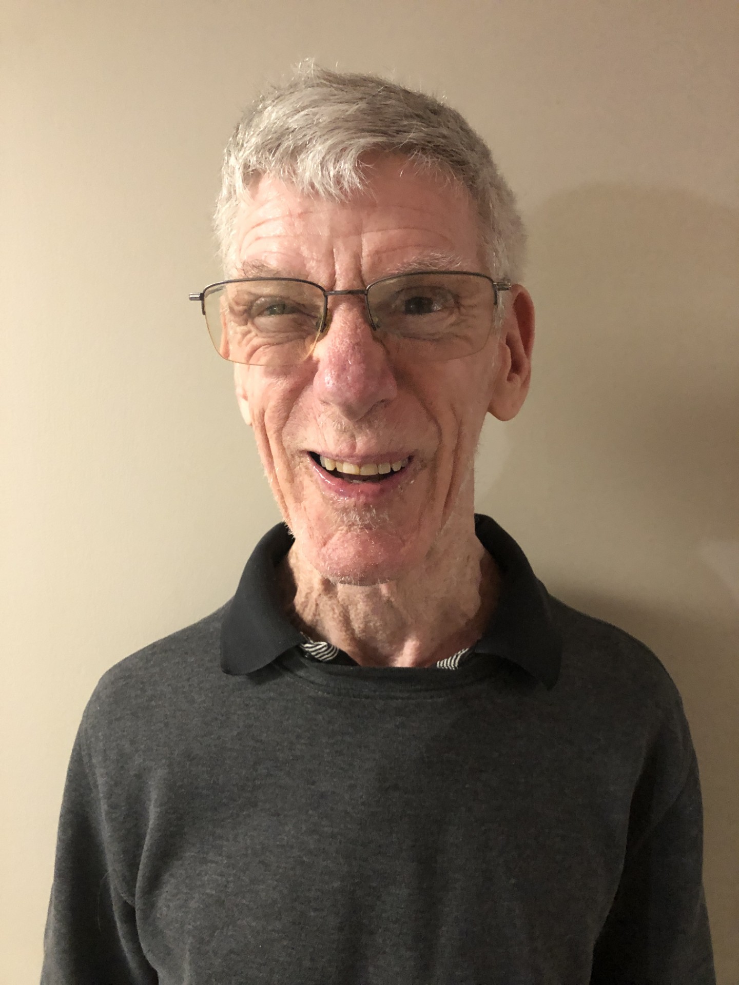 Brad smiles at the camera. He has light-toned skin, short, silver and white hair, and wears square glasses. He wears a black collared shirt.