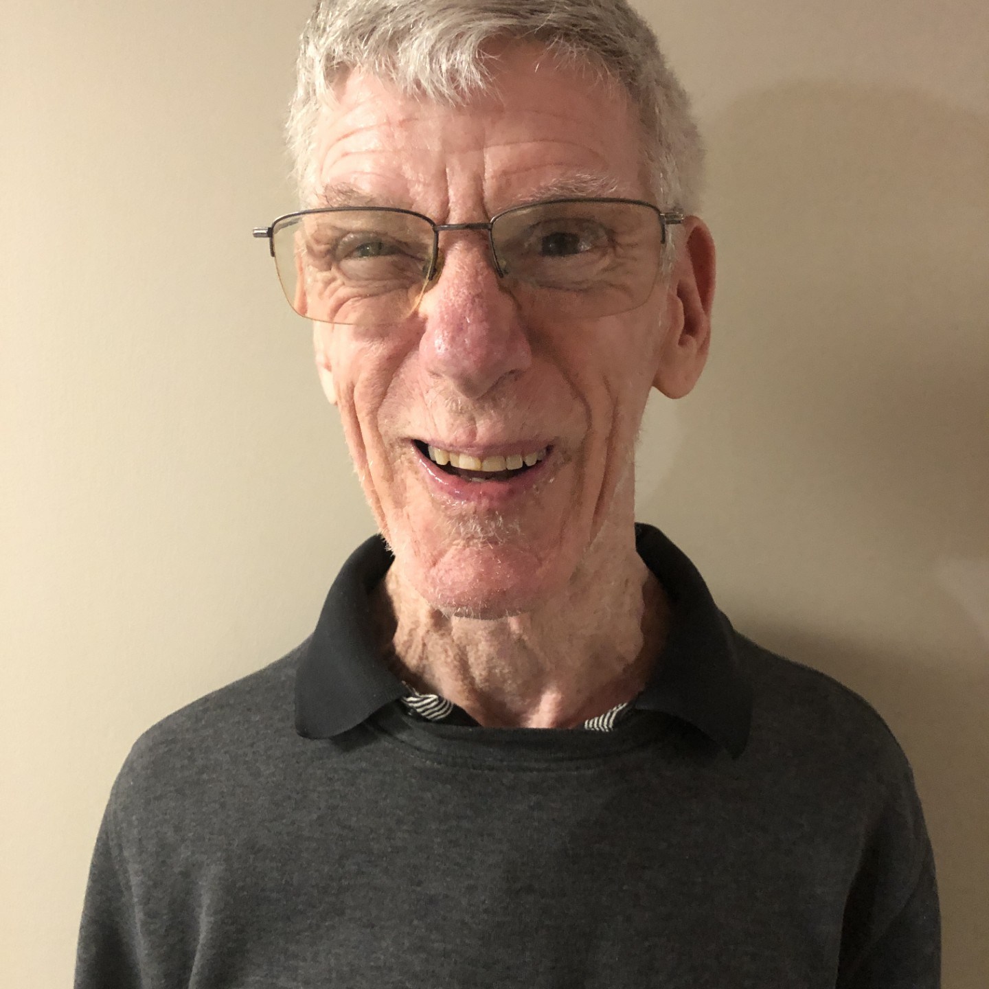 Brad smiles at the camera. He has light-toned skin, short, silver and white hair, and wears square glasses. He wears a black collared shirt.