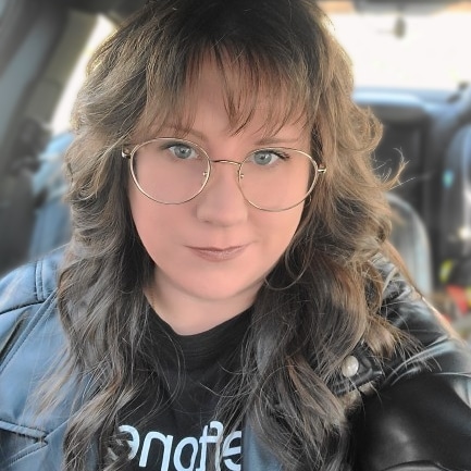 Meg smiles at the camera. She has light-toned skin, curled brown hair with bangs, and has large, round glasses. She wears a black leather jacket.