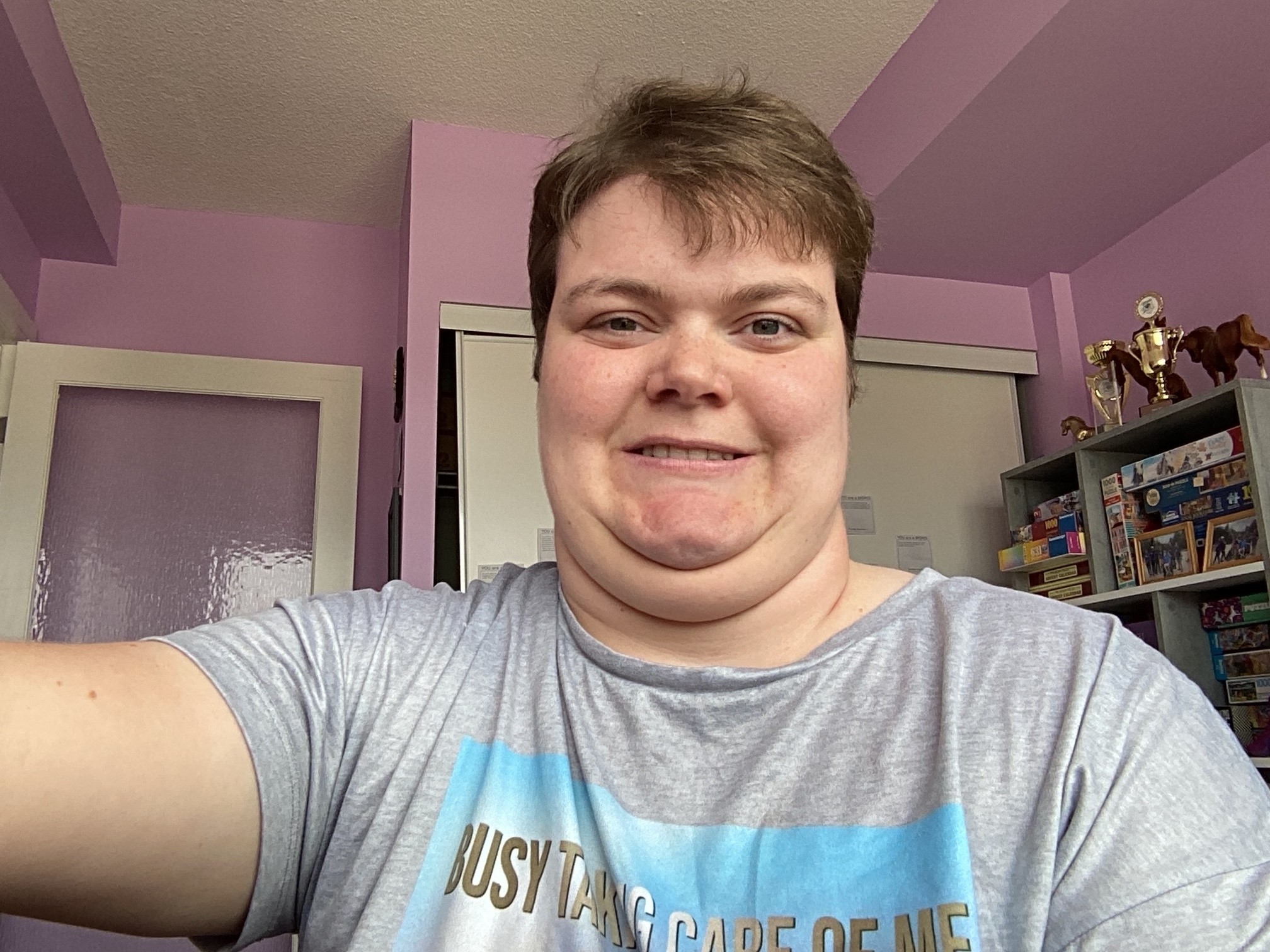 Maddy smiles happily at the camera. She has short, brown hair, light-toned skin, and wears a graphic t-shirt with grey and blue.