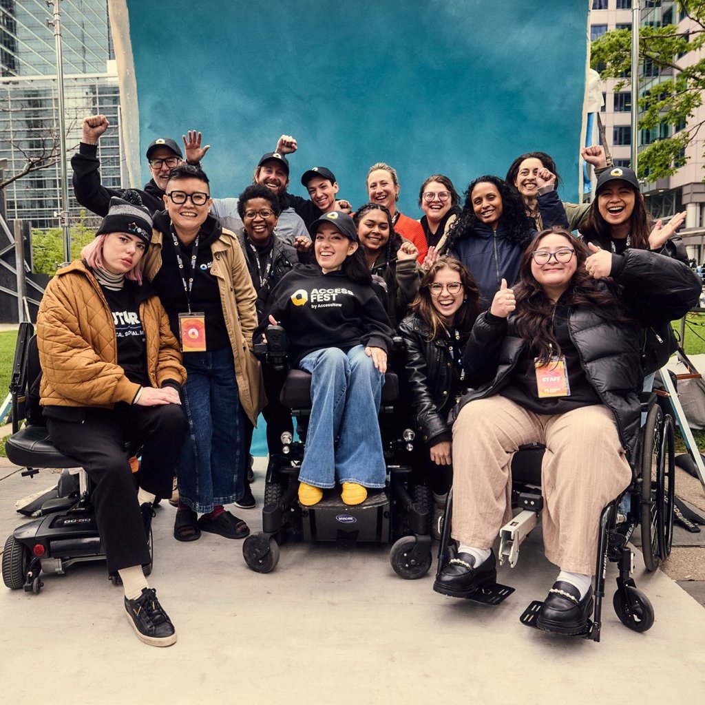 Group photo of the AccessNow team, including disabled and non-disabled individuals of diverse backgrounds, smiling and celebrating together in front of a bright blue backdrop outdoors. Several team members use wheelchairs or mobility devices