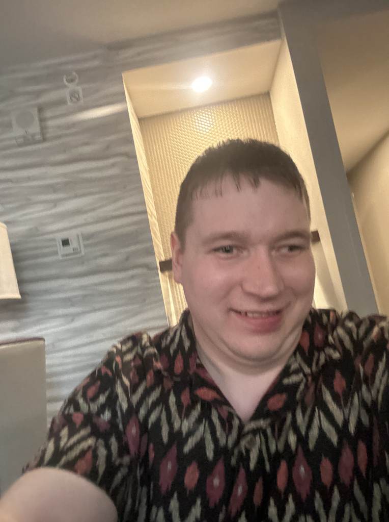 Justin smiles at the camera, he has short, brown hair, light-toned skin, and wears a geometric patterned shirt.