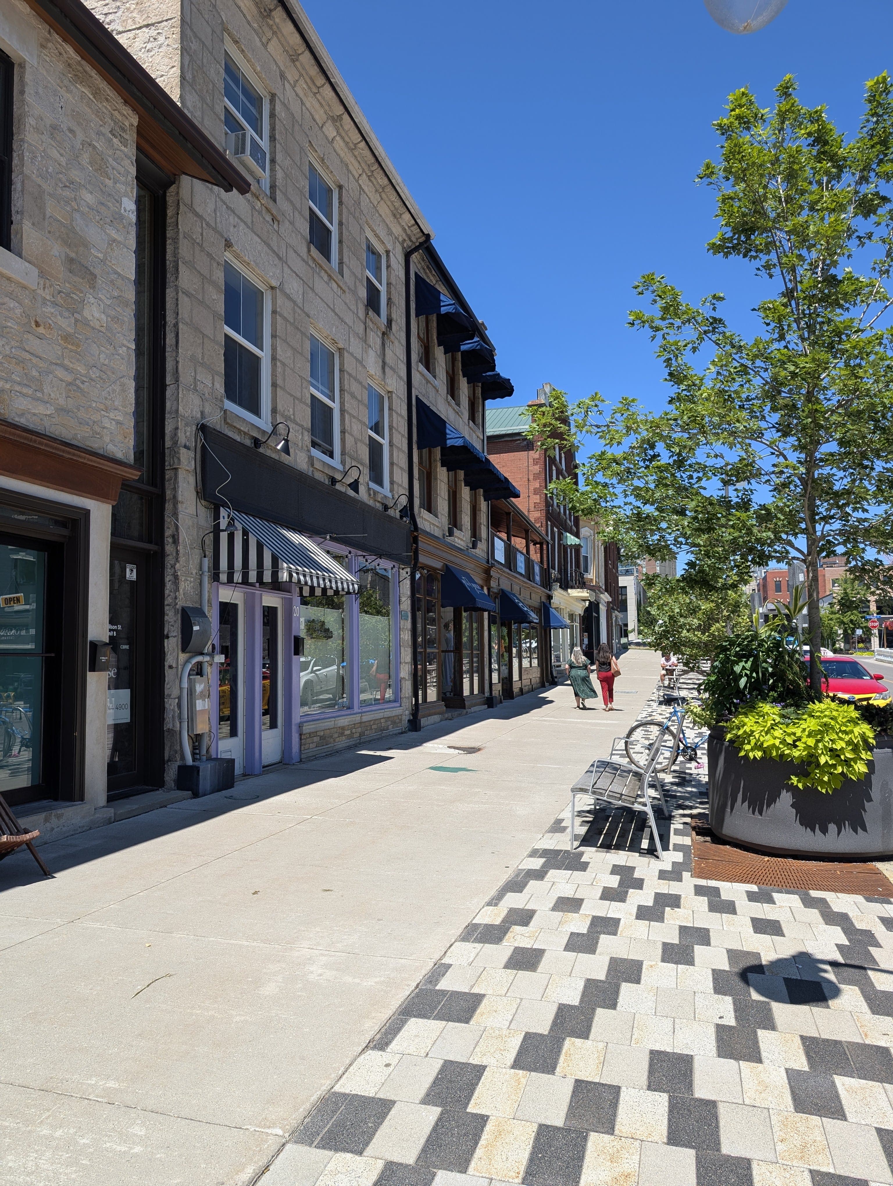 A row of row of Downtown Guelph businesses line the sidewalk with trees, benches, and bikes. Two women walk by the businesses in the distance.