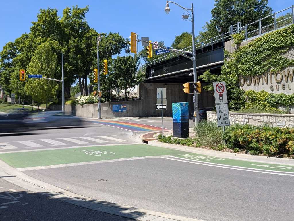 A busy street intersection with bright green bike lanes, and a vibrant rainbow crosswalk lead into Downtown Guelph, marked by a plant-covered, stone wall.