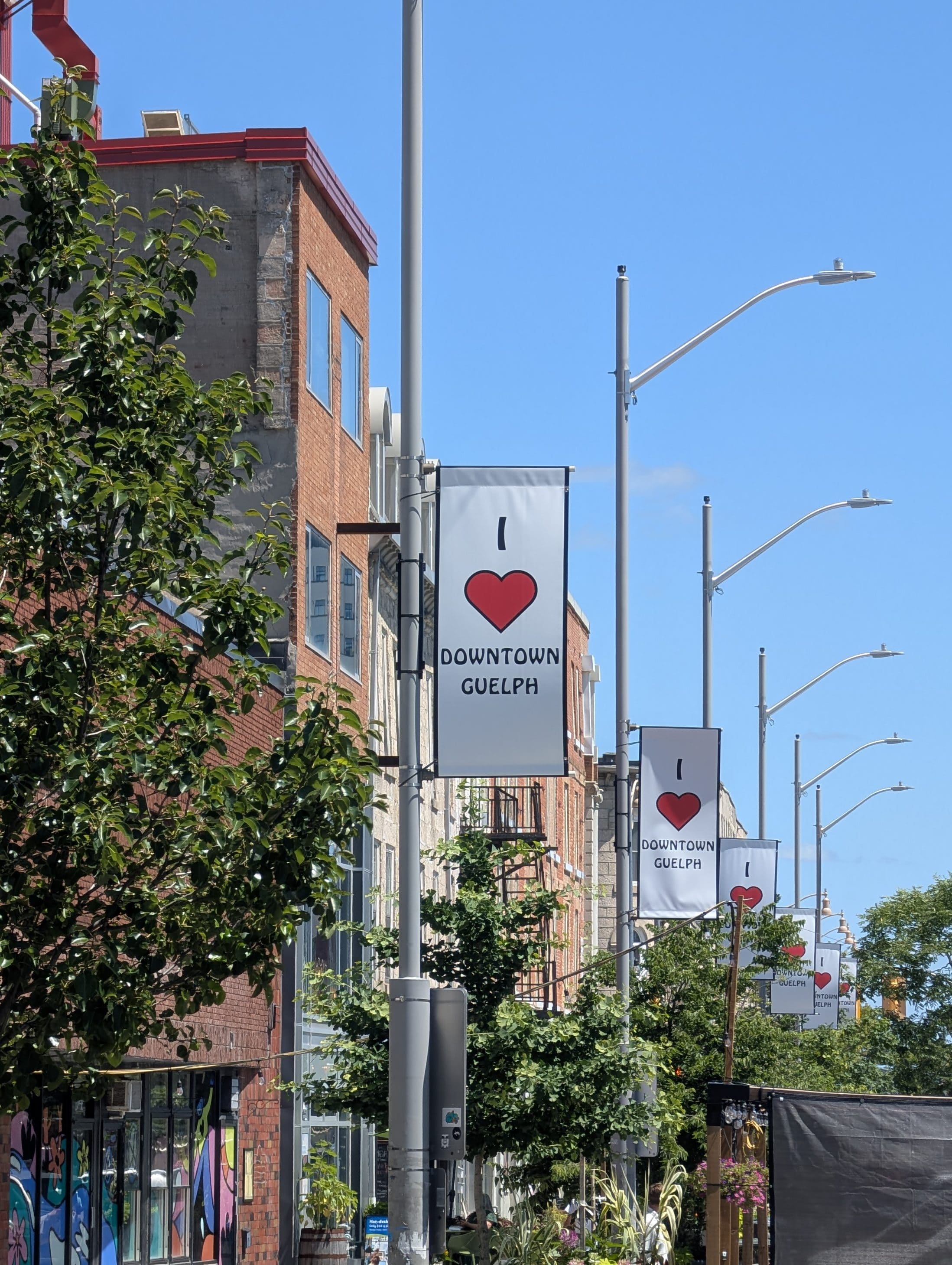 A row of "I heart Downtown Guelph" banners on streetlights line the sidewalk with brick buildings and trees under a clear blue sky.