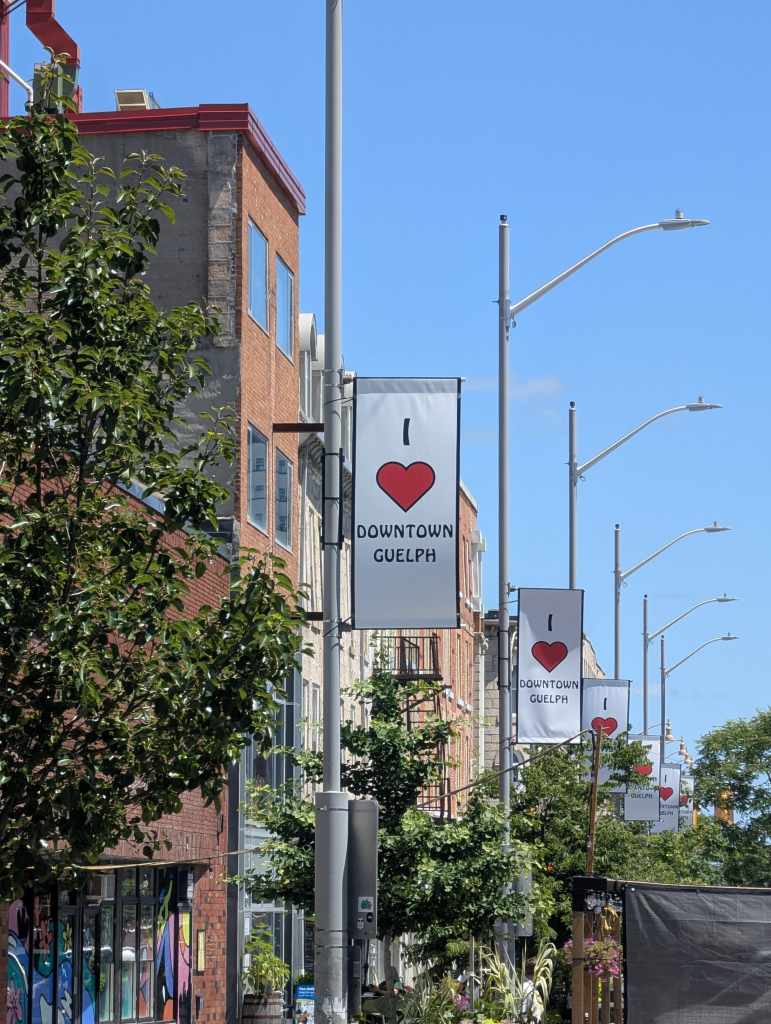 A row of "I heart Downtown Guelph" banners on streetlights line the sidewalk with brick buildings and trees under a clear blue sky.