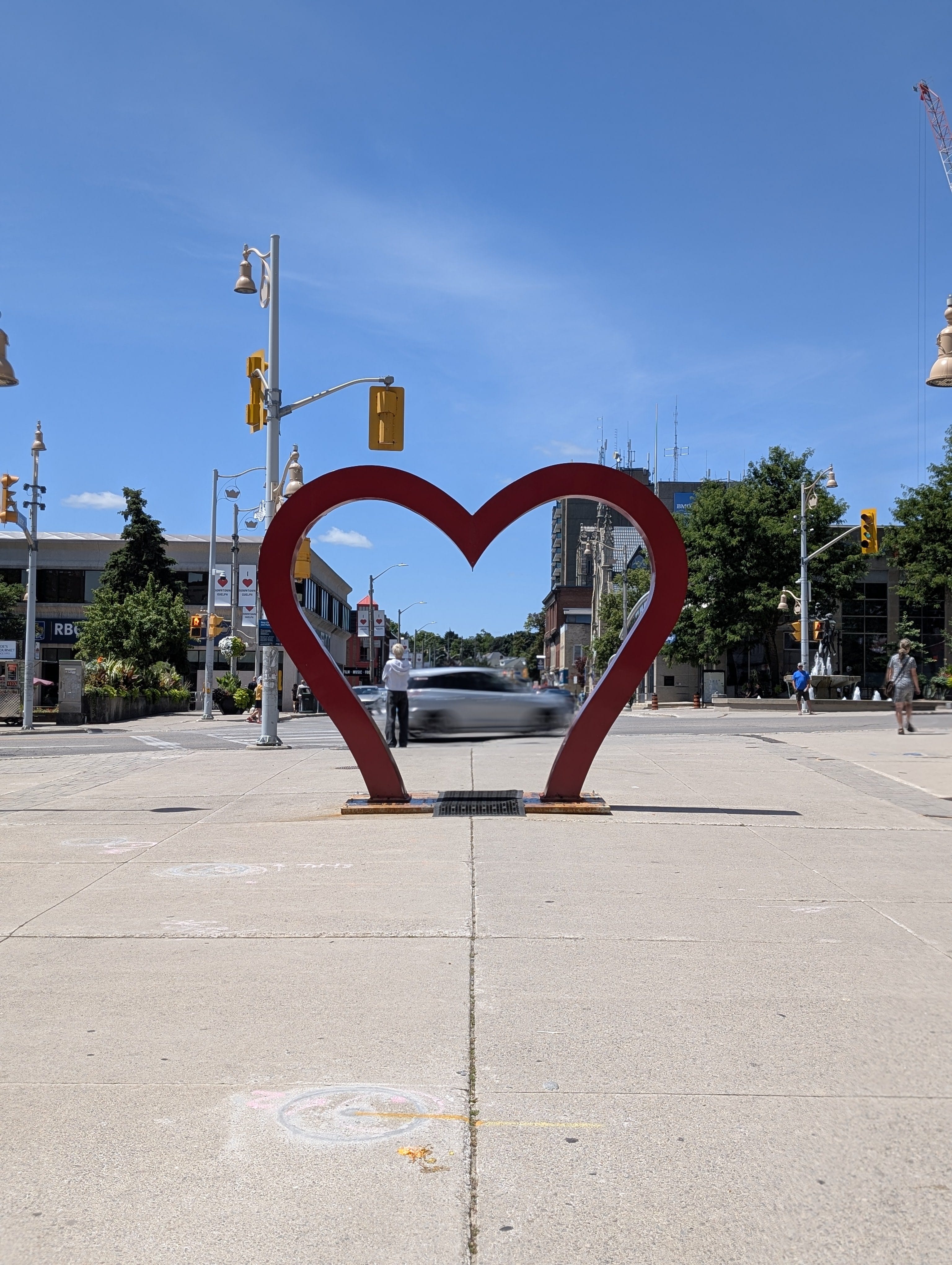A red heart-shaped sculpture is in focus on a Guelph street. The sky is clear and blue on a bright, sunny day.