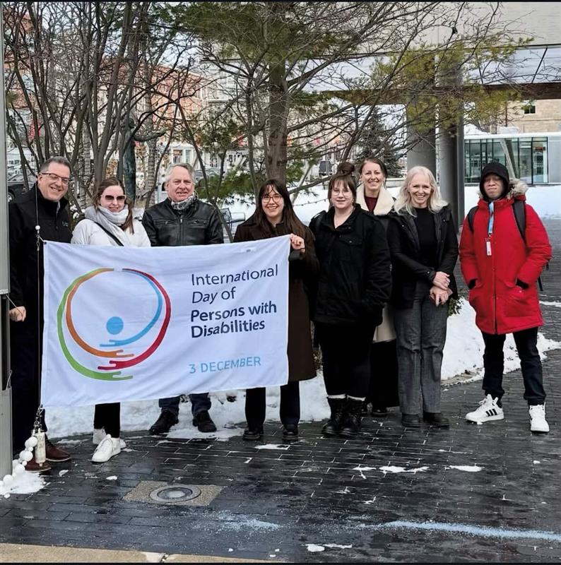Mayor Cam Guthrie, city councilors, city staff and local participants stand outside city hall holding an International Day of Persons with Disabilities flag.